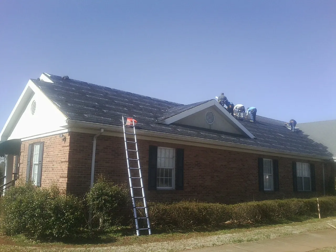 Crew tearing shingles off a brick ranch roof with a ladder set — Metal Roof Repair work by our crew in Crescent
